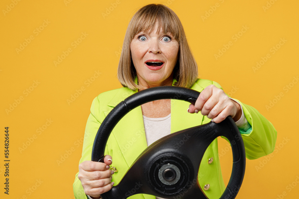 Elderly confused sad woman 50s years old wearing green jacket white t-shirt hold steering wheel driving car look camera isolated on plain yellow background studio portrait. People lifestyle concept. Elderly confused sad woman 50s years old wearing green jacket white t-shirt hold steering wheel driving car look camera isolated on plain yellow background studio portrait. People lifestyle concept.