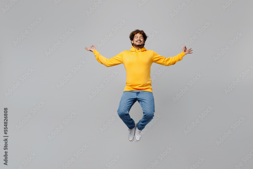 Full body young Indian man 20s he wearing casual yellow hoody jump high hold spreading hands in yoga om aum gesture relax meditate try to calm down isolated on plain grey background studio portrait