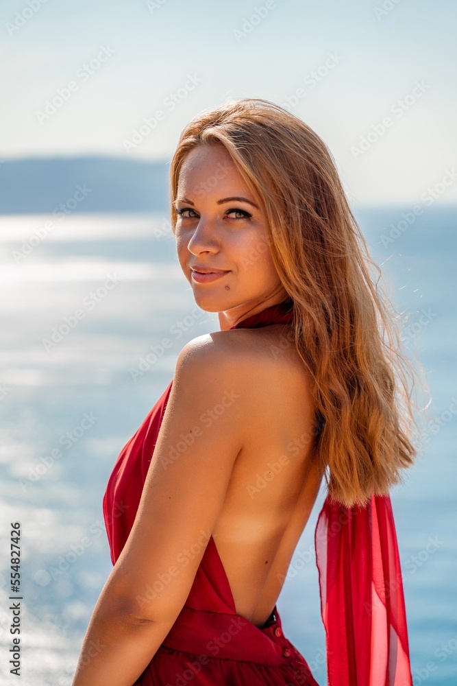 Naklejka premium Smiling young woman in a red dress looks at the camera. A beautiful tanned girl enjoys her summer holidays at the sea. Portrait of a stylish carefree woman laughing at the ocean.