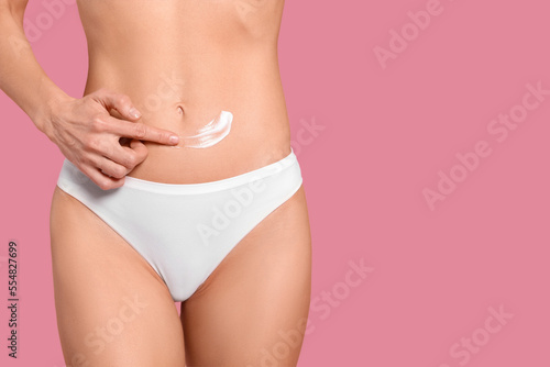 Woman applying body cream onto her belly against pink background, closeup. Space for text
