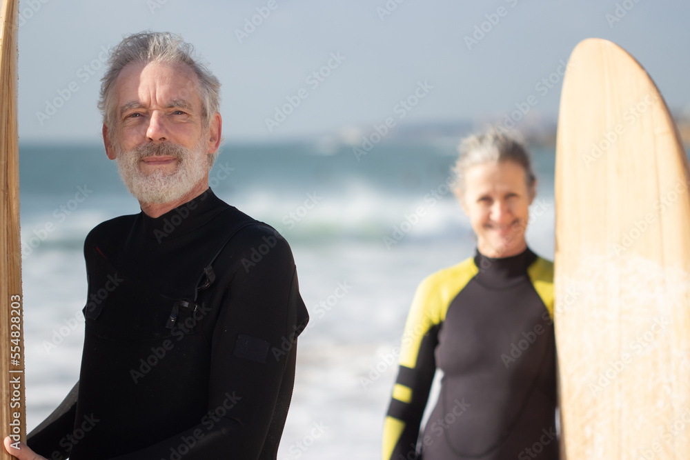 Portrait of aged couple training surfing in summer. Smiling man and woman behind him standing on sea shore holding wooden boards, looking at camera. Sport activity and relations of aged people concept