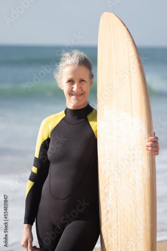 Portrait of happy senior woman preparing for surfing in sea. Attractive woman in wetsuit standing with board on sandy beach near sea and looking at camara. Sport and health of aged people concept