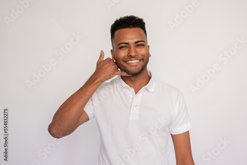 Portrait of happy young man making call gesture over white background. Latin American guy wearing white T-shirt looking at camera and smiling. Contact and advertising concept