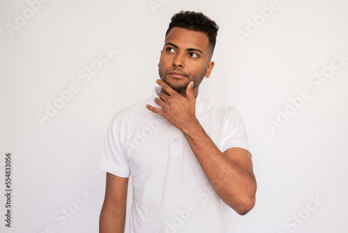 Portrait of young man looking away with pensive expression. Latin American guy wearing white T-shirt posing with hand on chin over white background. Thinking and planning concept