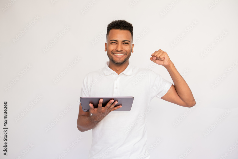 Cheerful young man using digital tablet. Male Indian model with brown eyes and curly hair in casual clothes using pad technology device with face of winner. Success, achievement concept Cheerful young man using digital tablet. Male Indian model with brown eyes and curly hair in casual clothes using pad technology device with face of winner. Success, achievement concept