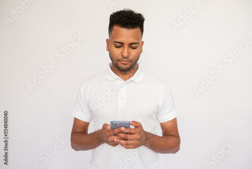 Concentrated young man using smartphone in his hands. Male Indian model with brown eyes and curly hair in casual clothes looking at mobile screen reading message or news. Modern technology concept