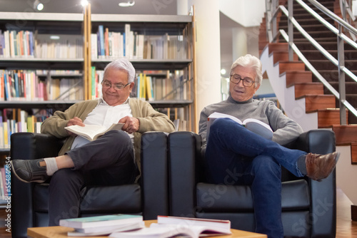 Positive aged men reading together. Two gray-haired students enjoying studying in university library finding information about technologies and discussing it. Studying of adult people concept