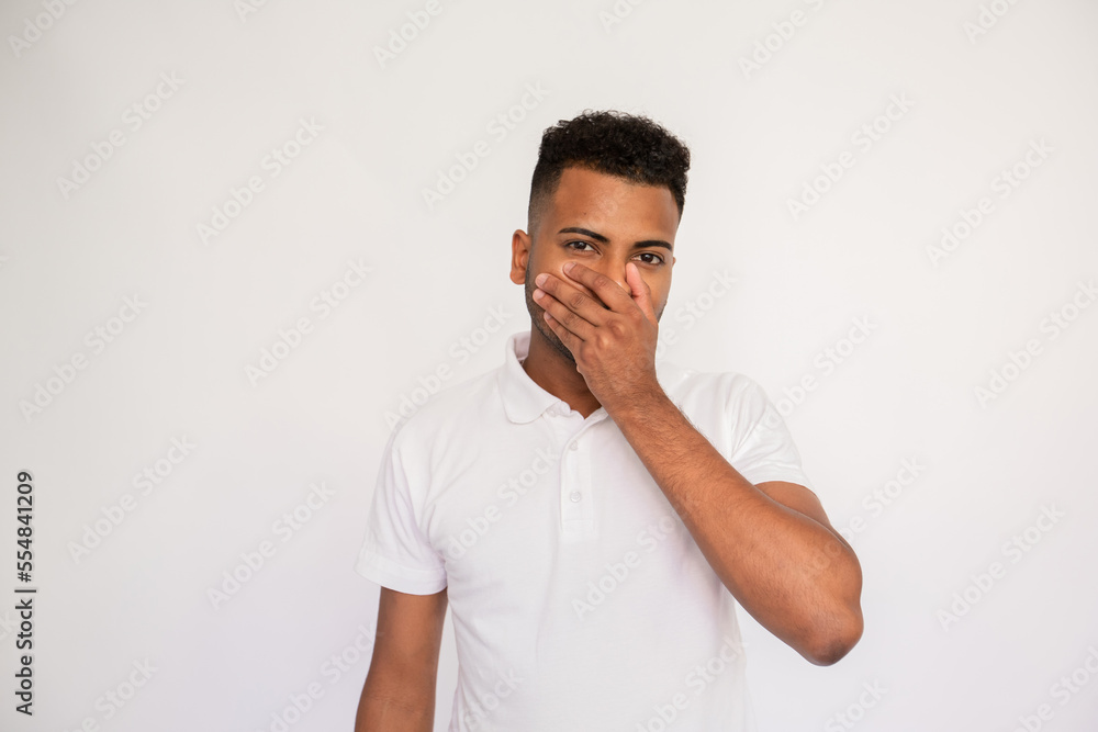 Serious young man covering his mouth with hand. Male Indian model with brown eyes and curly hair in white polo shirt standing in stupor, closing his mouth hearing shocking news. Surprise concept Serious young man covering his mouth with hand. Male Indian model with brown eyes and curly hair in white polo shirt standing in stupor, closing his mouth hearing shocking news. Surprise concept