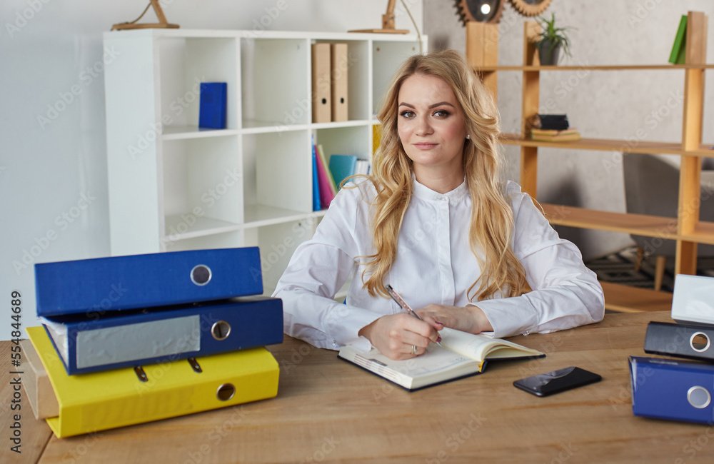 beautiful caucasian business woman sitting indoors, writing notes in notebook. stylish pretty female in shirt sits at table, works, studies with copybook. work, education, job beautiful caucasian business woman sitting indoors, writing notes in notebook. stylish pretty female in shirt sits at table, works, studies with copybook. work, education, job