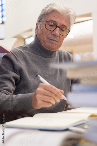 Portrait of senior man preparing for classes in library. Serious gray-haired man in glasses studying, reading books and writing down information with pen. Modern education for adult people concept