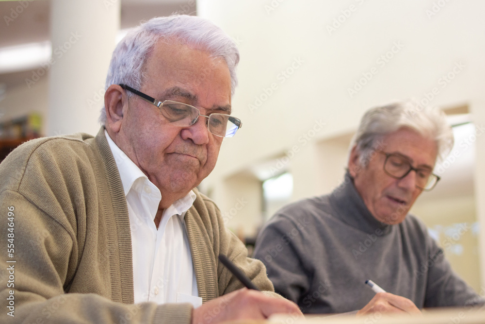 Serious aged men studying in library. Two gray-haired men in glasses sitting together at table reading about modern IT programs, making notes in notebooks. Adult people education concept Serious aged men studying in library. Two gray-haired men in glasses sitting together at table reading about modern IT programs, making notes in notebooks. Adult people education concept
