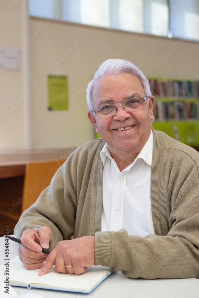 Portrait of aged man getting information from books. Happy Gray-haired man in glasses reading books about new technologies in library looking at camera and smiling. Education for adult people concept