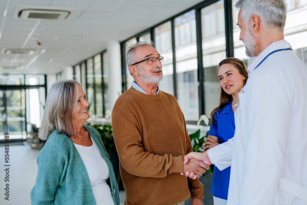 Mature doctor and his young colleague talking with his senior patients at hospital corridor. Mature doctor and his young colleague talking with his senior patients at hospital corridor.