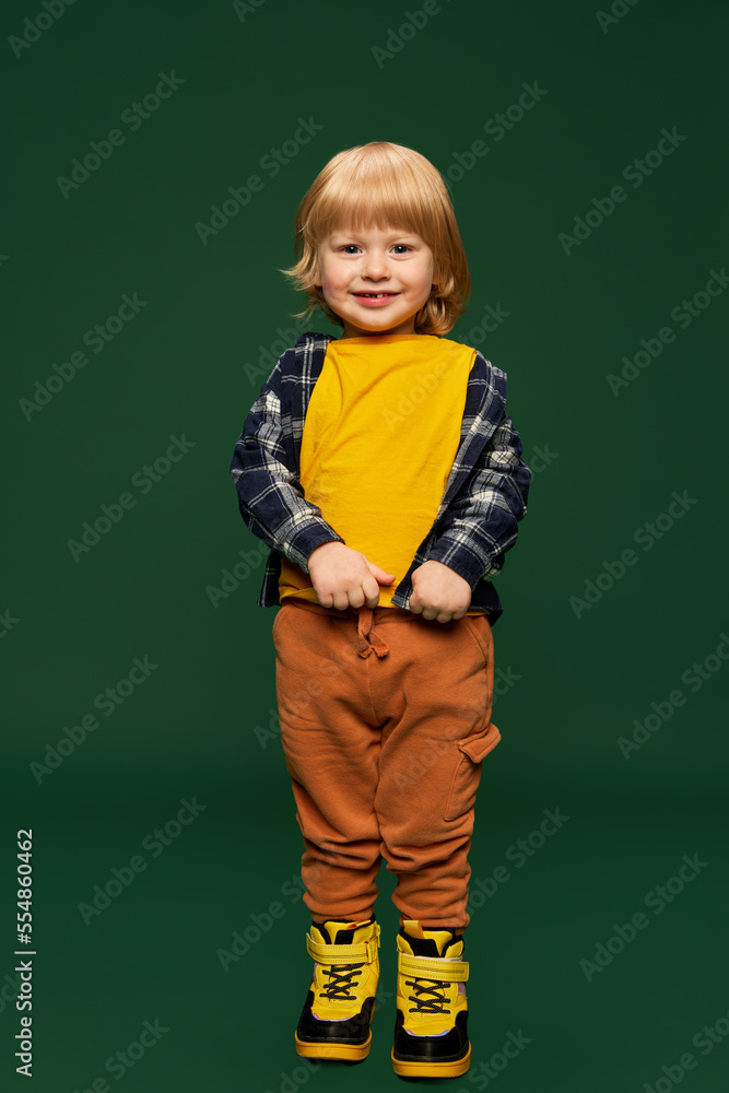 Portrait of adorable little boy, child in fashionable casual clothes posing with smile over green studio background. Concept of childhood, emotions Portrait of adorable little boy, child in fashionable casual clothes posing with smile over green studio background. Concept of childhood, emotions