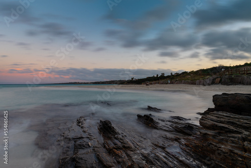 sunset over the picturesque white sand beach and turquoise waters at La Pelosa Beach with rocks in the foreground