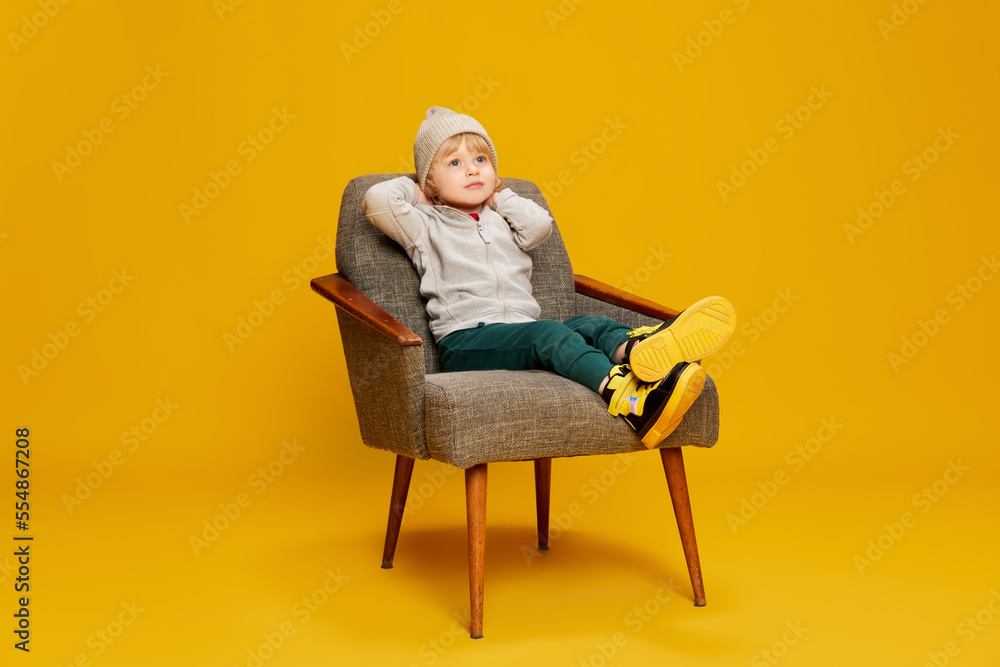 Portrait of little boy, child posing with dreamy look, sitting on chair over yellow studio background. Relaxation. Concept of childhood, emotions Portrait of little boy, child posing with dreamy look, sitting on chair over yellow studio background. Relaxation. Concept of childhood, emotions