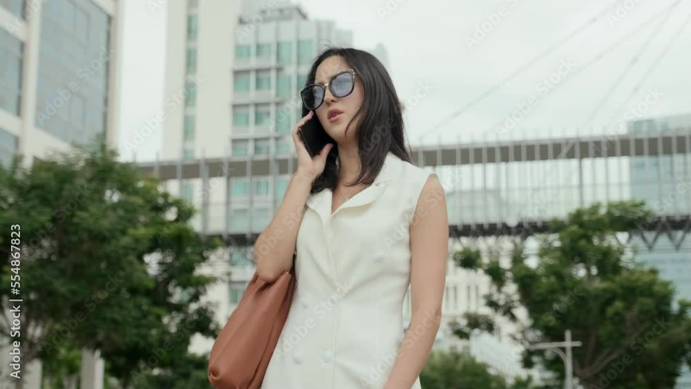 Young elegant businesswoman in white dress with brown leather handbag on shoulder talking on mobile phone while standing in front of camera in urban environment on summer day Young elegant businesswoman in white dress with brown leather handbag on shoulder talking on mobile phone while standing in front of camera in urban environment on summer day