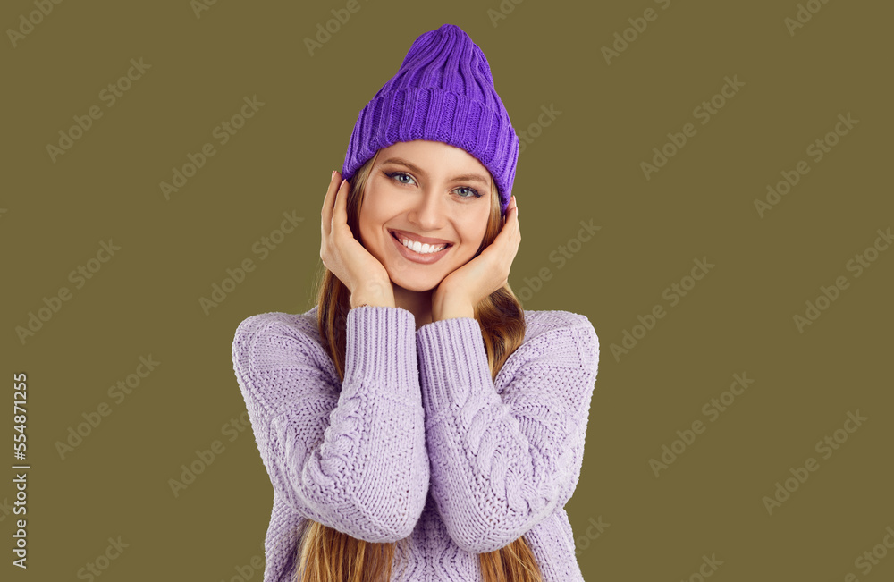 Portrait of beautiful woman in cozy winter clothes. Happy young fashion model in warm purple knitted hat and lavender sweater standing isolated on dark green background, smiling and looking at camera Portrait of beautiful woman in cozy winter clothes. Happy young fashion model in warm purple knitted hat and lavender sweater standing isolated on dark green background, smiling and looking at camera