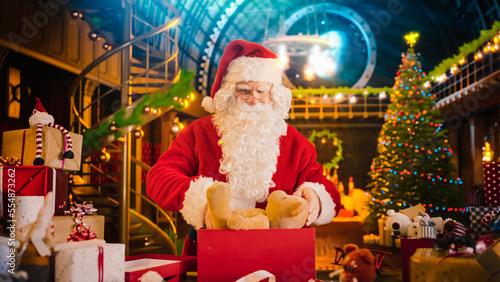 Santa Claus in Studio Workshop: Wrapping and Packing Christmas Gifts for all the Good Children to be Delivered on the Magical New Year Eve. He Prepares the Teddy Bear As A Presents for an Excited Kid photo