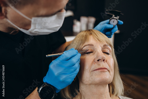 Charming older woman getting face lift shot over light studio background. Unrecognizable beautician or plastic surgeon making beauty injection to attractive senior female patient