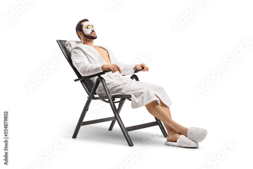 Man in a bathrobe with a face mask and cucumber on eyes sitting on a chair