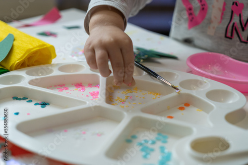 a child's hand sorts shiny colored pieces for decoration in a white paint palette