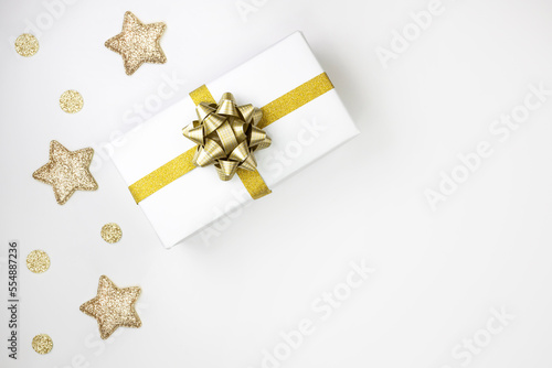 Gift box in white paper with a gold bow on a white background with gold stars. Flat lay, place for text.