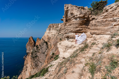 A beautiful young woman in a white light dress with long legs stands on the edge of a cliff above the sea waving a white long dress, against the background of the blue sky and the sea.