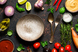 Cooking food background. Fresh vegetables, spices and herbs with plate, cutlery and utensils on black table