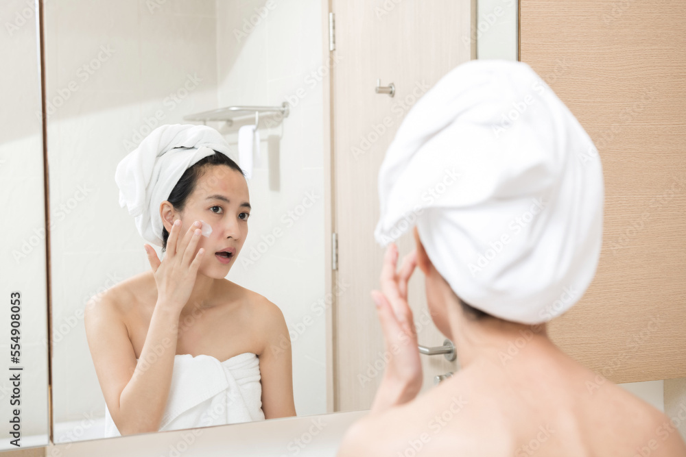 Happy young Asian woman applying face lotions while wearing a towel and touching her face in bathroom Happy young Asian woman applying face lotions while wearing a towel and touching her face in bathroom