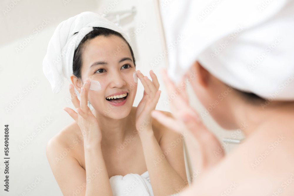 Happy young Asian woman applying face lotions while wearing a towel and touching her face in bathroom