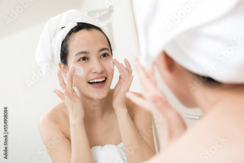 Happy young Asian woman applying face lotions while wearing a towel and touching her face in bathroom