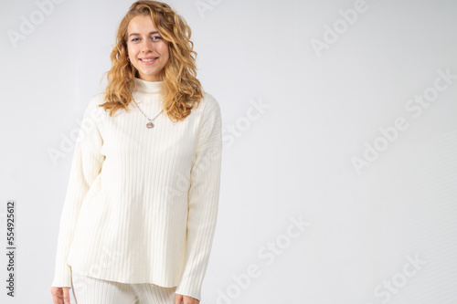 A young blonde girl with curly hair stands and smiles on a white background. The model is dressed in a white knitted suit
