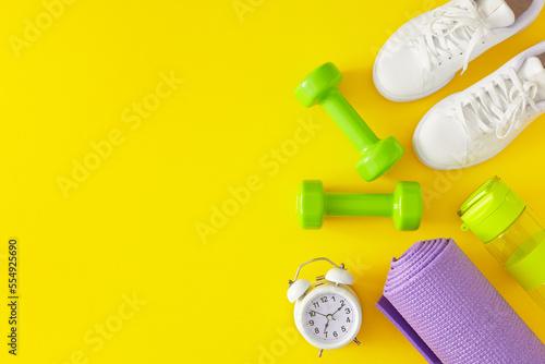 Active living concept. Flat lay photo of green dumbbells, white sports shoes, bottle of water, violet exercise mat and alarm clock on yellow background with copy space. Minimal fitness idea.