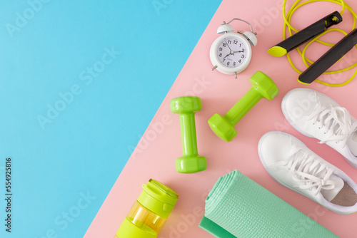 Active living concept. Flat lay composition of green dumbbells, white sneakers, exercise mat, skipping rope, bottle of water, alarm clock on pastel pink and blue background with copy space. 