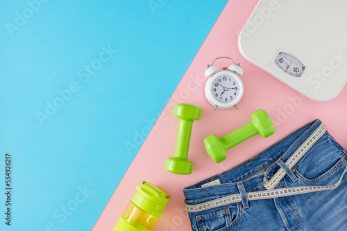 Slimming concept. Top view photo of blue jeans with tape measure, dumbbells, scales and alarm clock on pink and blue background. Flat lay with copy space.