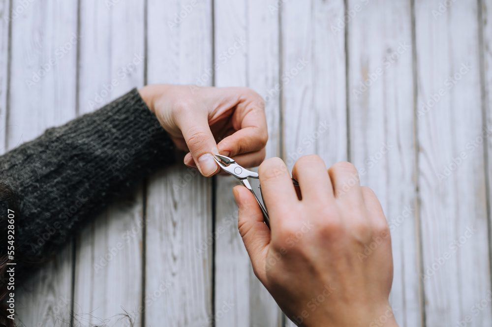 Obraz premium Hands of a woman, a girl close-up, making herself a manicure, cutting cuticles with special scissors on fingers, nails on a wooden white background. Close-up photography, work, beauty, art.