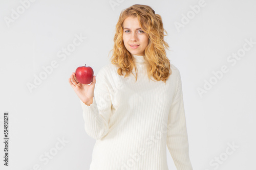 A pretty young blonde woman with curly hair holds a red ripe apple in her hand. The girl is dressed in a white warm knitted suit, she stands on a white background