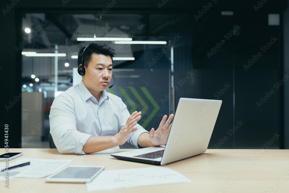 Online sales. Telemarket. A young Asian salesman is sitting in a headset in the office at a table in front of a laptop. Sells goods, accepts orders, advises, customer support hotline. Online sales. Telemarket. A young Asian salesman is sitting in a headset in the office at a table in front of a laptop. Sells goods, accepts orders, advises, customer support hotline.