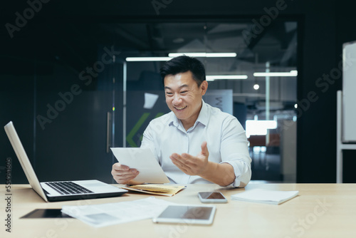 A young man, an Asian businessman, sits smiling in the office at the table, works with documents, holds papers, contracts, bills in his hands.