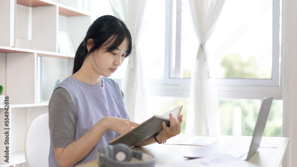 Young female student reading a book in a private office, Long distance education, Textbooks from books, Find out more, In the office there is a computer or laptop placed on the table, Study. Young female student reading a book in a private office, Long distance education, Textbooks from books, Find out more, In the office there is a computer or laptop placed on the table, Study.