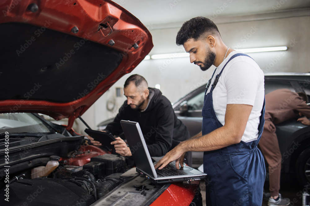 Two young male mechanic using laptop and tablet, recording automobile engine checking collecting detailed information during their work on car workshop. Service maintenance during engine repair. Two young male mechanic using laptop and tablet, recording automobile engine checking collecting detailed information during their work on car workshop. Service maintenance during engine repair.