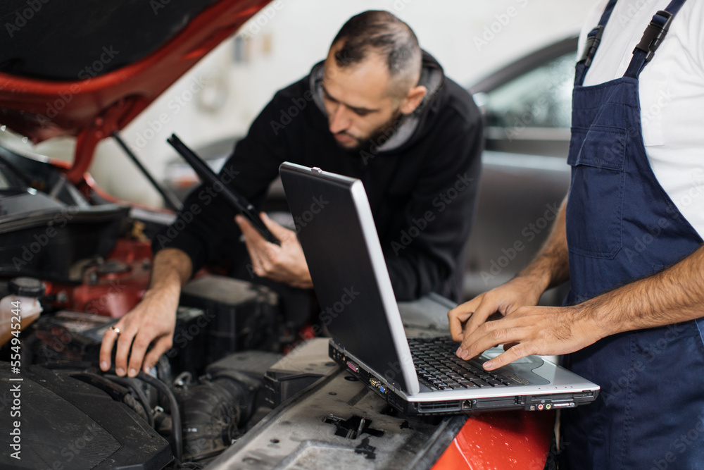 Close up view of hand of young male mechanic using laptop and his colleague with tablet, recording automobile engine checking collecting detailed information during their work on car workshop. Close up view of hand of young male mechanic using laptop and his colleague with tablet, recording automobile engine checking collecting detailed information during their work on car workshop.