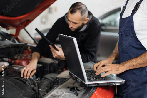 Close up view of hand of young male mechanic using laptop and his colleague with tablet, recording automobile engine checking collecting detailed information during their work on car workshop.