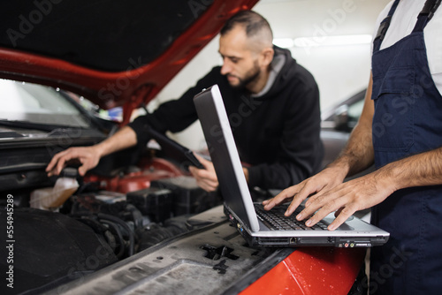 Close up view of hand of young male mechanic using laptop and his colleague with tablet, recording automobile engine checking collecting detailed information during his work on car workshop. © sofiko14