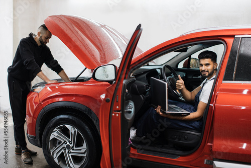 Young male mechanic using laptop sitting inside red car and his colleague with tablet under hood recording automobile engine checking collecting detailed information during their work showing thumb up