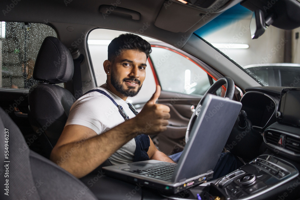 Young bearded man car technician mechanic sitting inside red car repairing problem of engine, during system checking detail, using laptop computer for maintenance and fixing in garage showing thumb up Young bearded man car technician mechanic sitting inside red car repairing problem of engine, during system checking detail, using laptop computer for maintenance and fixing in garage showing thumb up