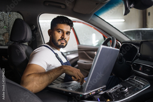 Young bearded man car technician mechanic sitting inside red car repairing problem of engine, during system checking detail, using laptop computer for maintenance and fixing in garage. © sofiko14