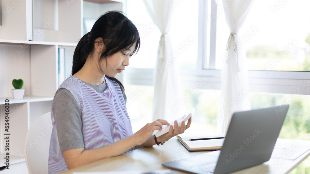 Asian woman working at home, Press the calculator to calculate the income - expenses of the house and use the laptop to record the information and check the accuracy, Home lifestyle. Asian woman working at home, Press the calculator to calculate the income - expenses of the house and use the laptop to record the information and check the accuracy, Home lifestyle.