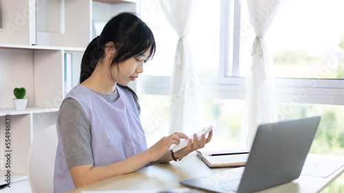 Asian woman working at home, Press the calculator to calculate the income - expenses of the house and use the laptop to record the information and check the accuracy, Home lifestyle.